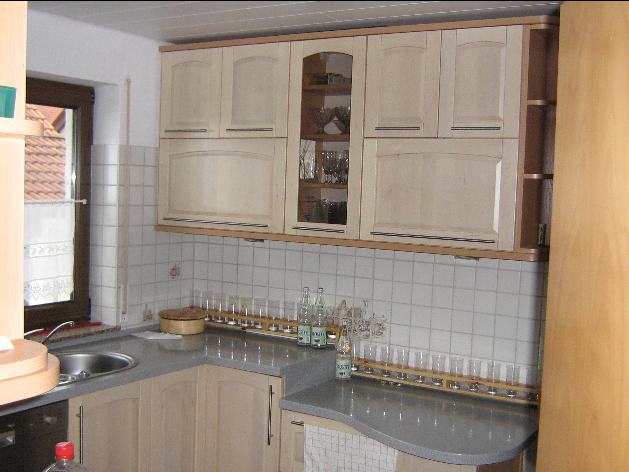 Jasna kuchnia z kremowymi szafkami, szklankami na blacie i oknem z widokiem na dach.