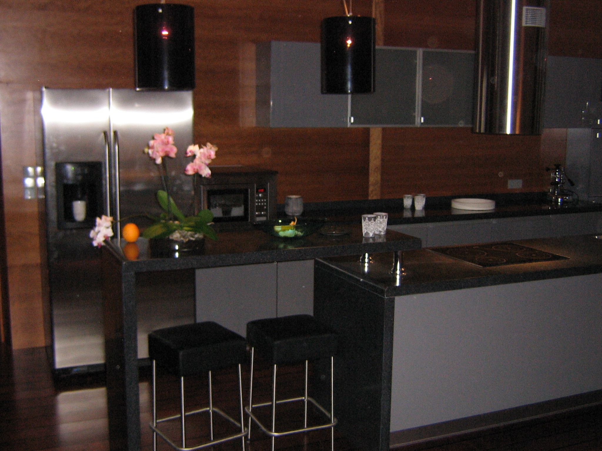 Nowoczesna kuchnia z ciemnymi blatami, stalową lodówką i wyspą z płytą indukcyjną, oświetlona wiszącymi lampami cylindrycznymi i ozdobiona kwiatami orchidei.