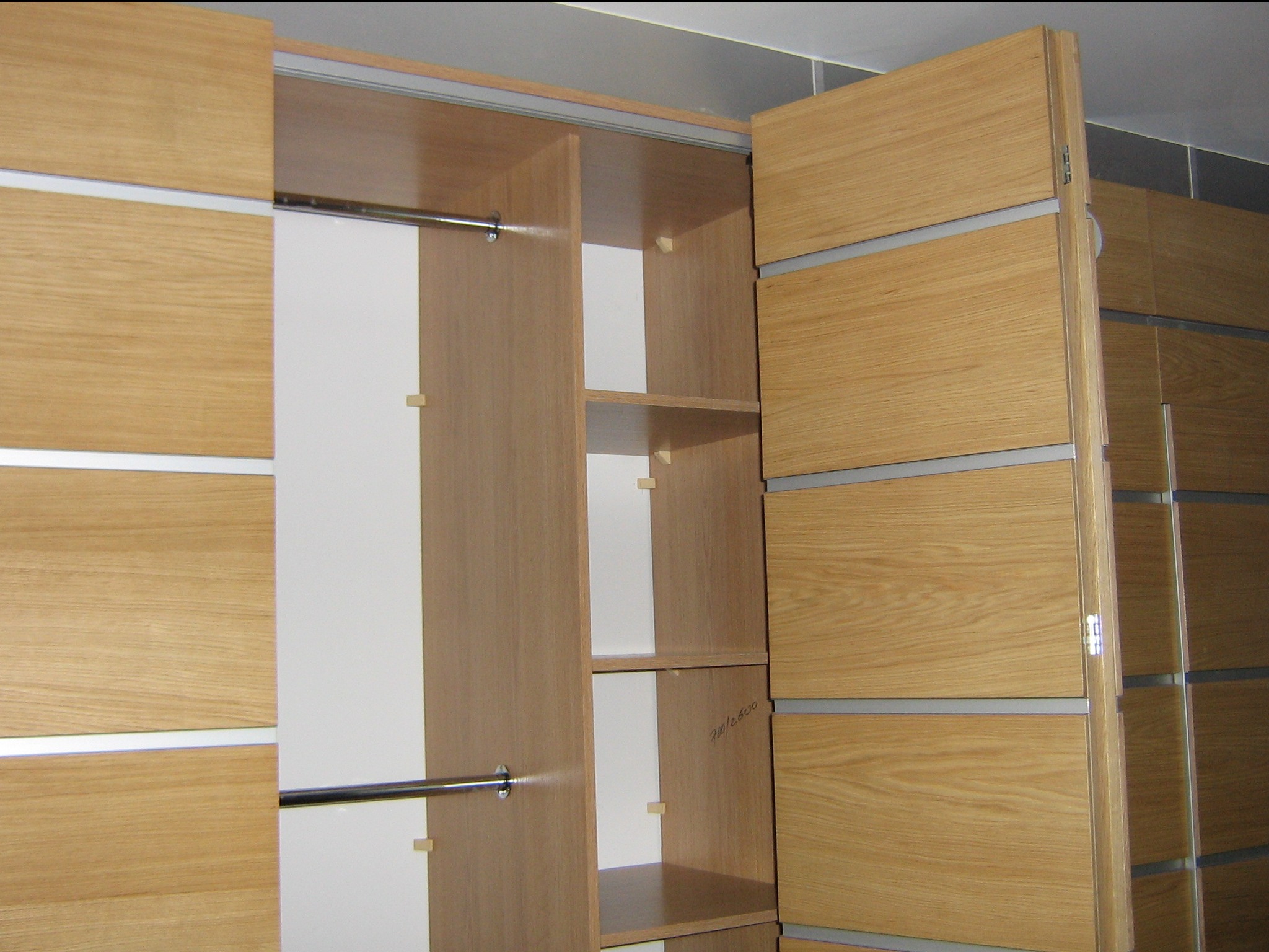 Otwarta szafa wnękowa z przesuwnymi drzwiami z jasnego drewna z poziomymi, metalowymi wstawkami, widoczne półki i drążek na ubrania, wnętrze szafy w kolorze jasnego drewna i bieli.