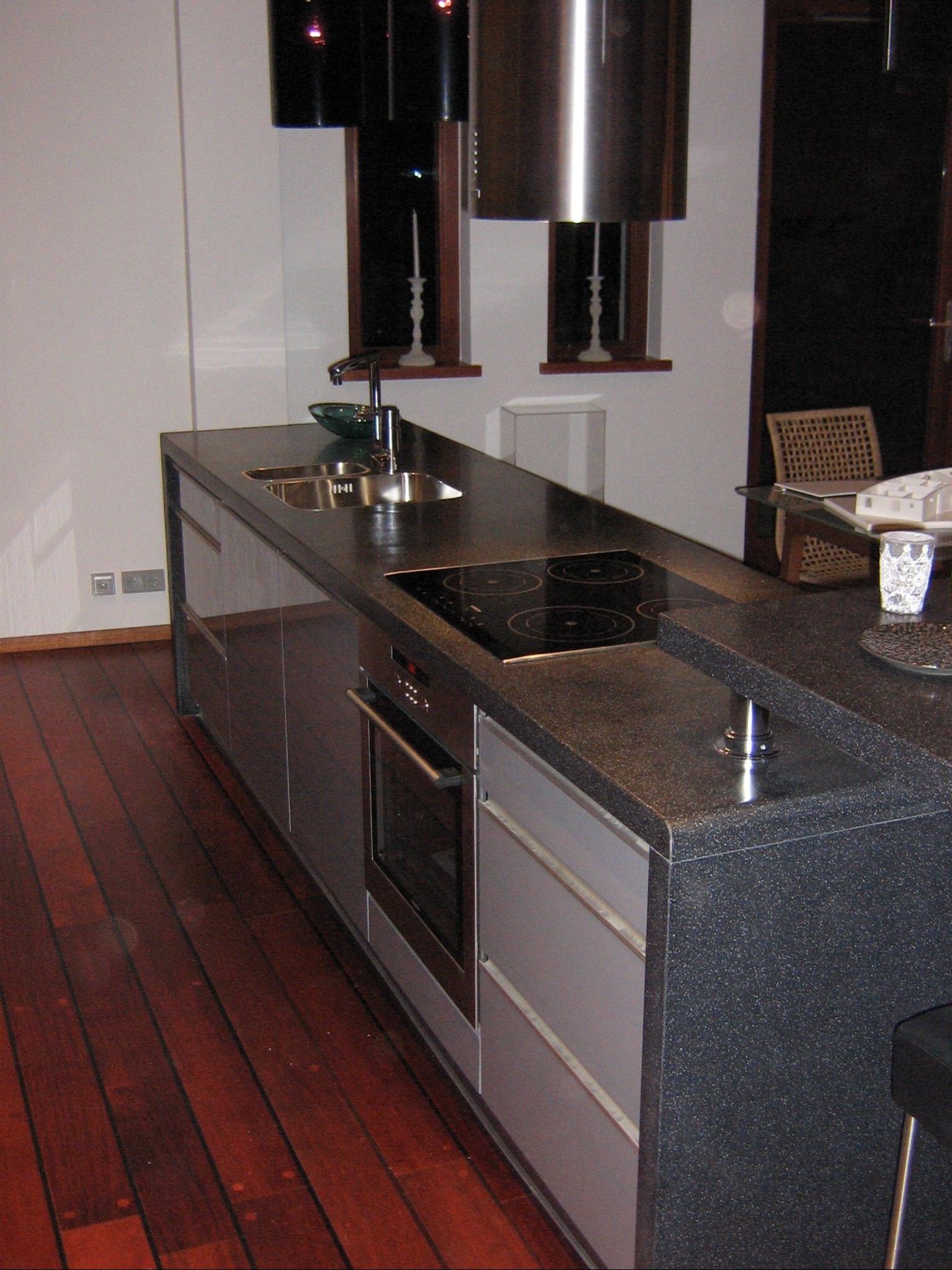 Nowoczesna wyspa kuchenna z ciemnym, teksturowanym blatem, wbudowaną płytą indukcyjną i piekarnikiem, uzupełniona drewnianą podłogą i minimalistycznym oświetleniem.