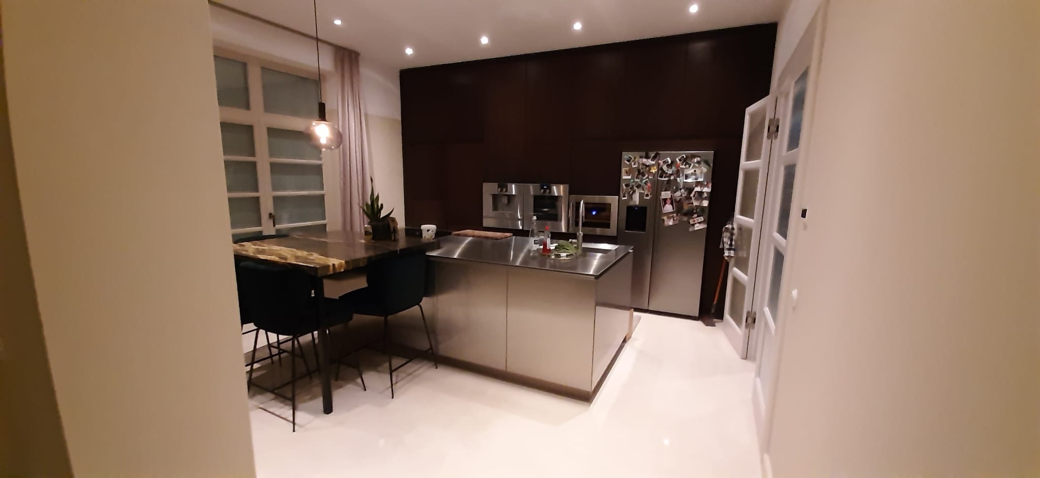 Nowoczesna kuchnia z wyspą ze stali nierdzewnej, ciemnobrązowymi szafkami i marmurowym stołem z krzesłami z ciemnej zieleni.