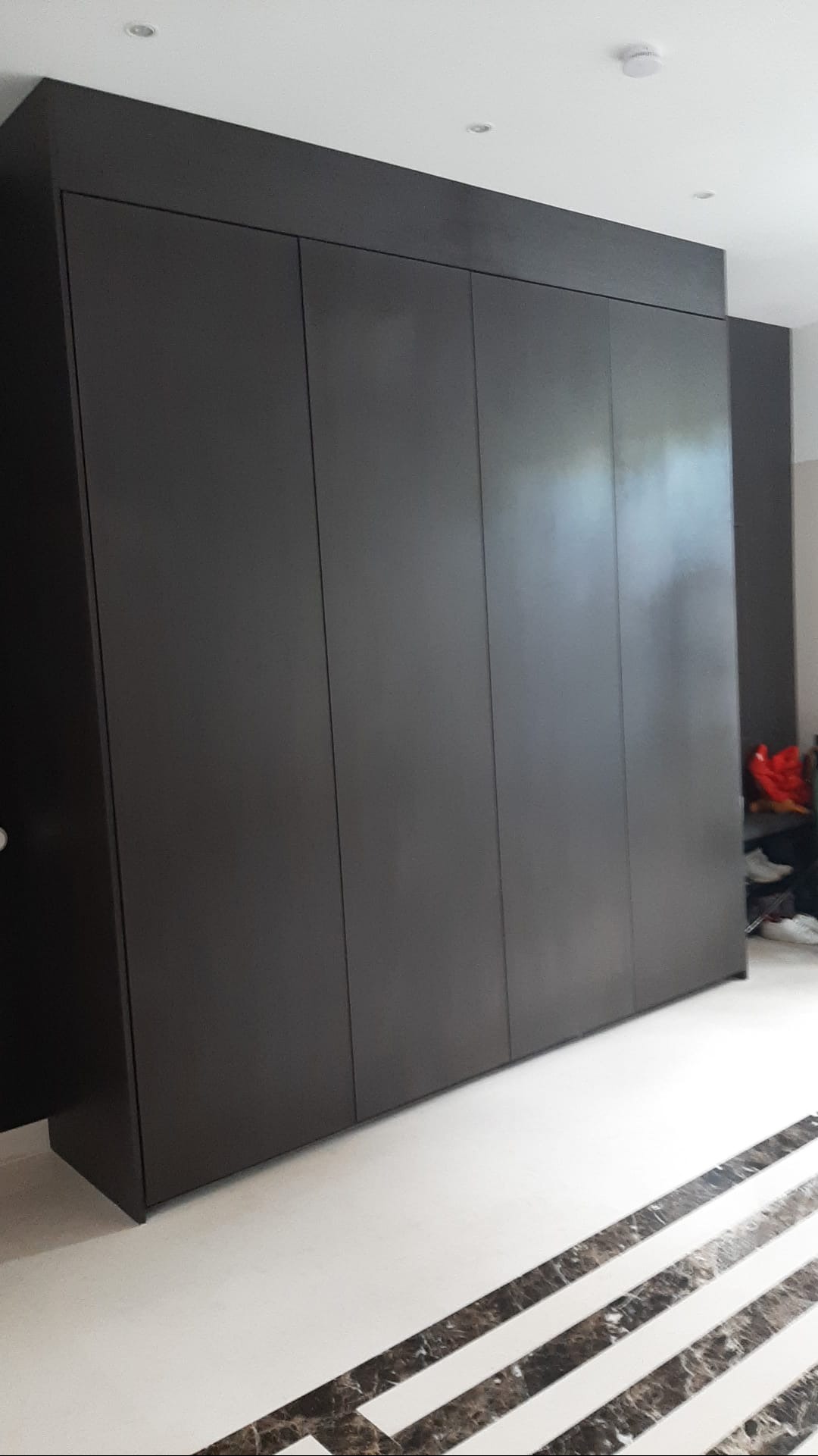Duża, matowa, ciemnoszara szafa wnękowa z czterema drzwiami, zabudowana od podłogi do sufitu, z widocznymi elementami wykończenia sufitu i ozdobną podłogą z marmurowym wzorem.