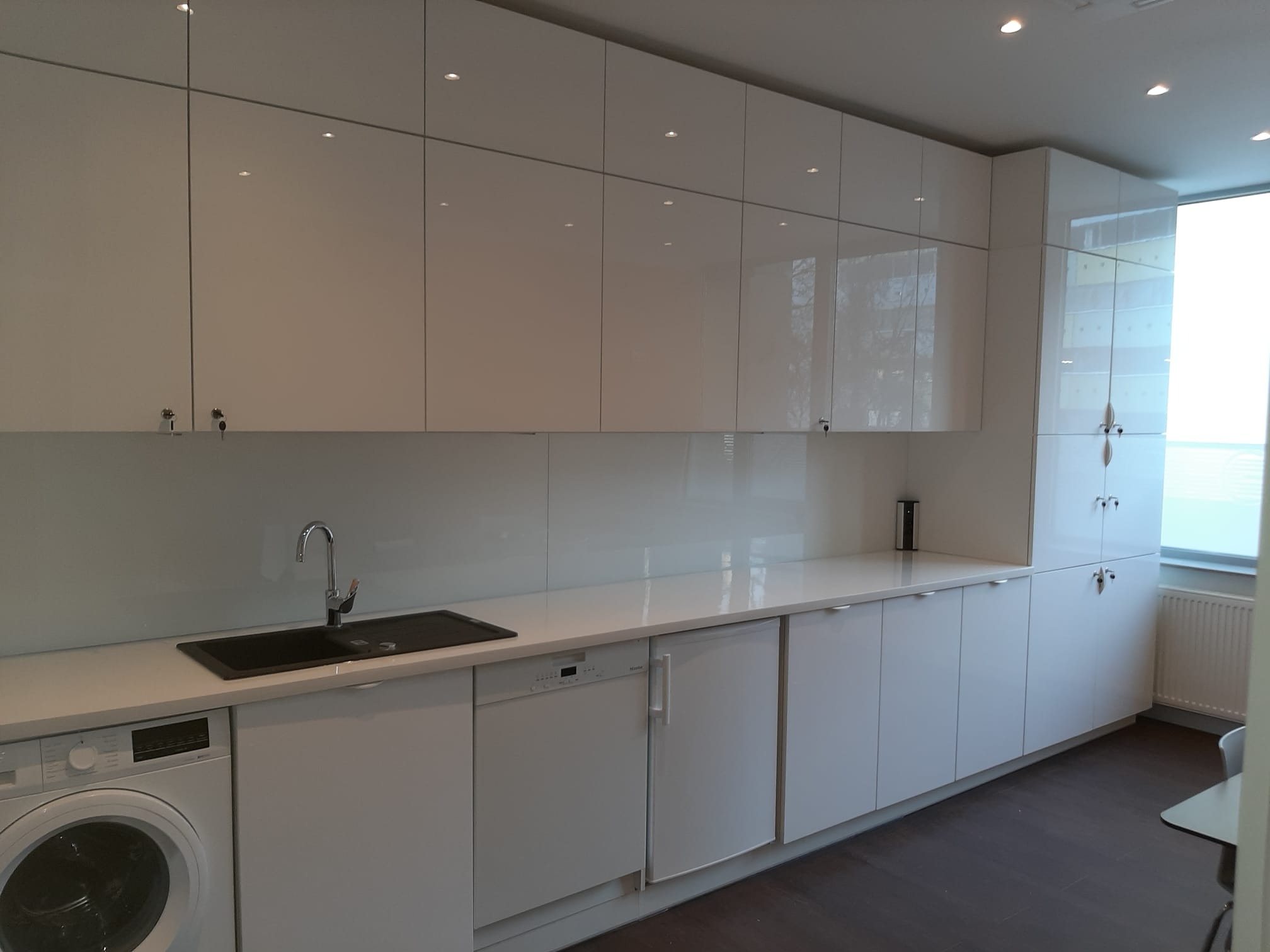 Biała, nowoczesna kuchnia z połyskującymi frontami szafek, czarnym zlewem i wbudowanymi sprzętami AGD, w tym pralką i zmywarką.