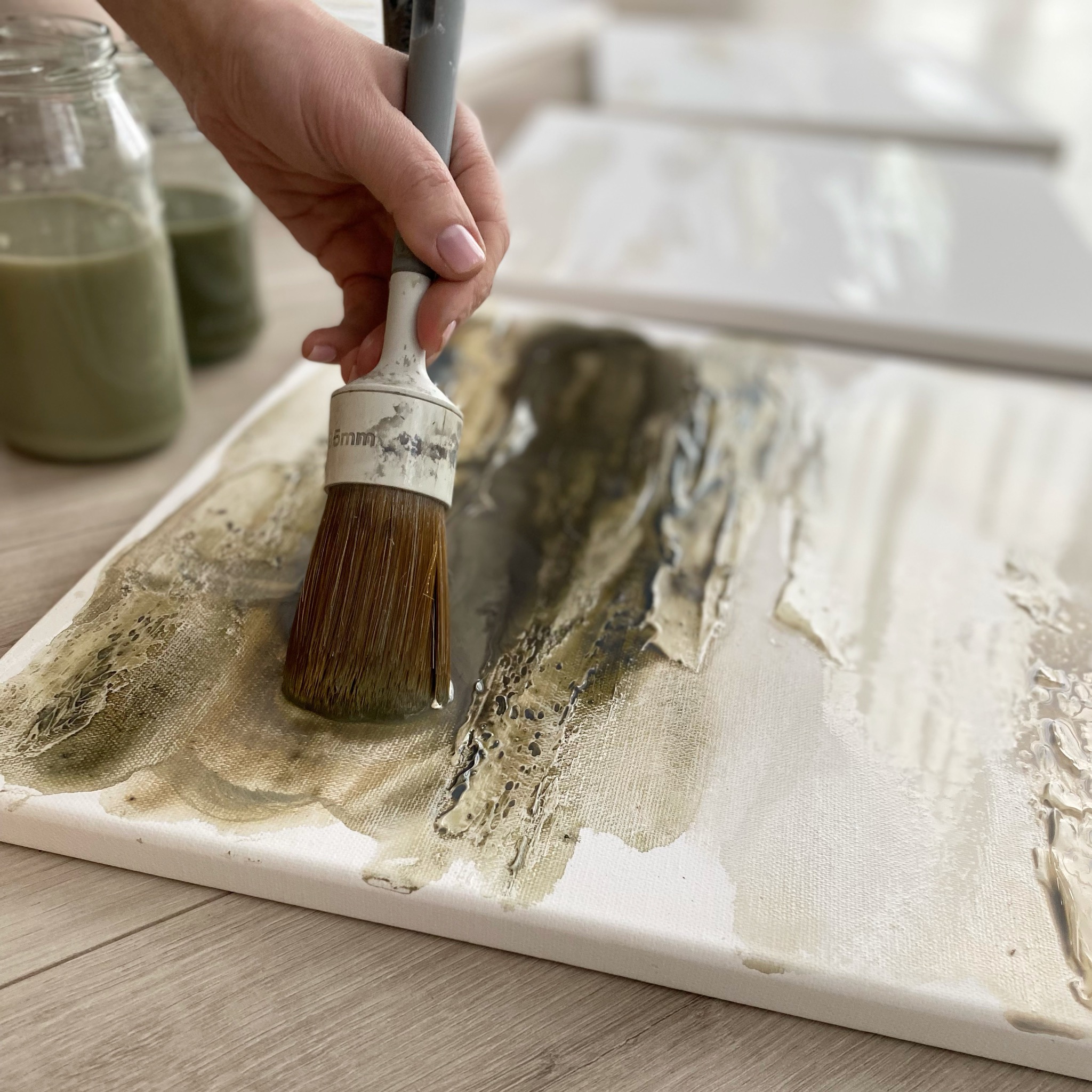 Artysta nakłada pędzlem farbę w odcieniach beżu i brązu na płótno, tworząc abstrakcyjną fakturę.