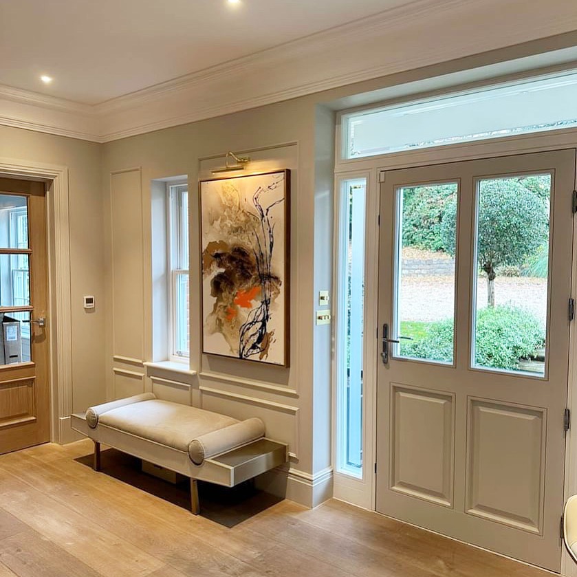 Abstrakcyjny obraz w brązowo-pomarańczowych tonacjach zawieszony w hallu nad beżową ławką, oświetlony kinkietem.