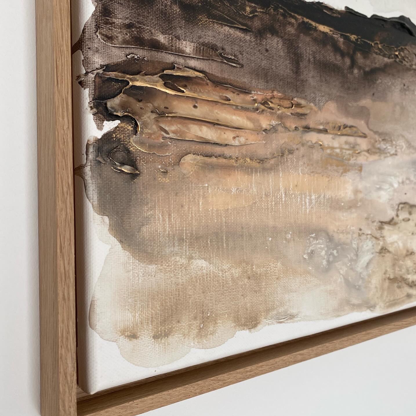 Abstrakcyjny obraz w odcieniach brązu i beżu z elementami złota, faktura z wyraźnymi, wypukłymi warstwami, rama z jasnego drewna, widok z boku.