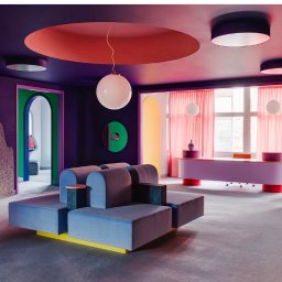 Olbud - Awangardowe wnętrze biura z geometrycznymi kształtami, fioletowymi ścianami, pastelowymi meblami i okrągłymi lampami wiszącymi.