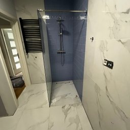 Olbud - Wykończona łazienka z kabiną prysznicową typu walk-in z niebieskimi płytkami, białe płytki imitujące marmur na ścianach i podłodze, widoczne niedokończone gniazdko elektryczne i grzejnik drabinkowy.