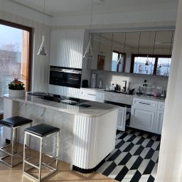 Olbud - Nowoczesna kuchnia z marmurowym blatem wyspowym, białymi szafkami z pionowymi żłobieniami, czarno-białą podłogą w geometryczny wzór i drewnianą podłogą w jodełkę, oświetlona wiszącymi lampami...