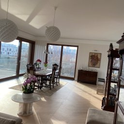 Olbud - Jasny salon z parkietem w jodełkę, dużymi oknami balkonowymi, okrągłym stołem z krzesłami, kwiatami w wazonach i dekoracyjnymi lampami wiszącymi.