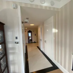 Wnętrze korytarza z kremowo-białymi tapetami w pionowe pasy, marmurową posadzką z czarnymi akcentami i drewnianą podłogą, widziane z perspektywy wejścia.