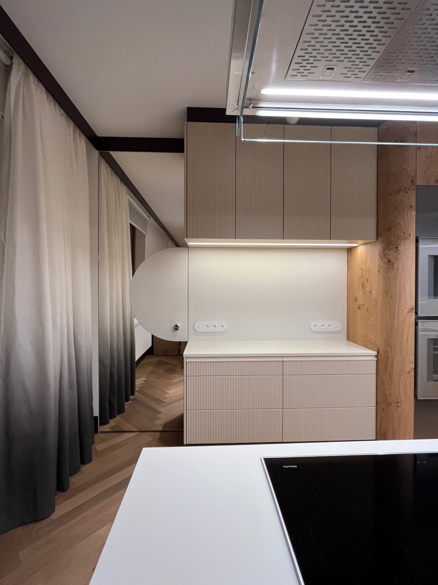 Nowoczesna kuchnia z beżowymi szafkami z ryflowanym frontem, białym blatem i drewnianym fornirem, widok na parkiet w jodełkę i zasłony ombre.