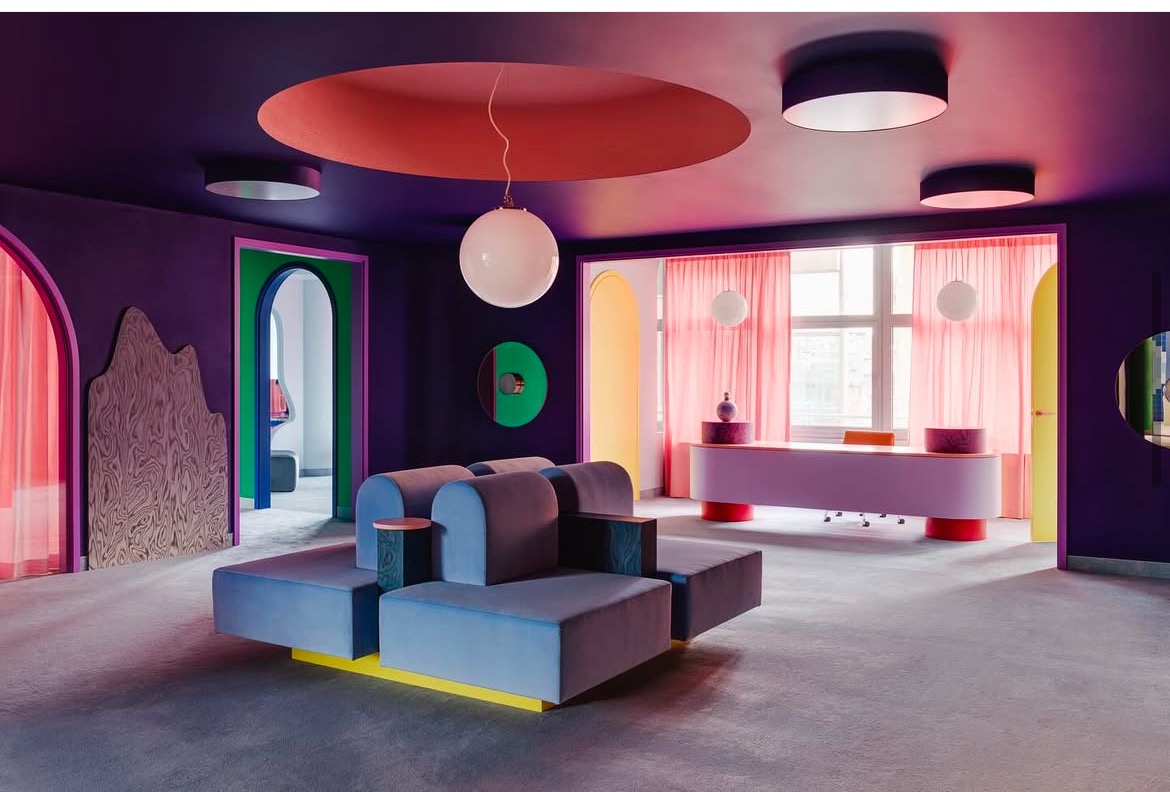 Awangardowe wnętrze biura z geometrycznymi kształtami, fioletowymi ścianami, pastelowymi meblami i okrągłymi lampami wiszącymi.