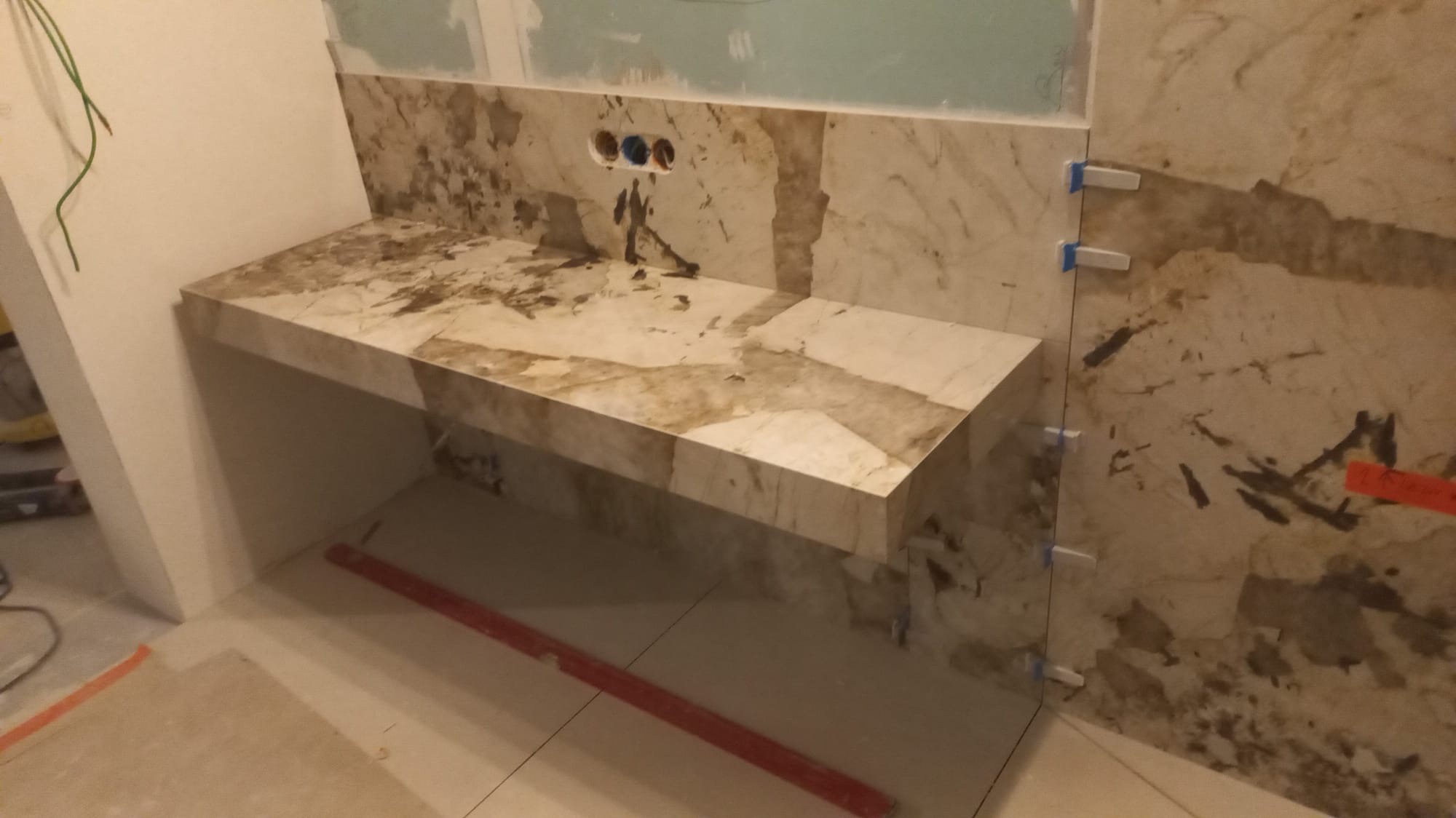 Montaż blatu z marmuru w łazience, widoczne płytki na ścianie z dystansami, instalacja elektryczna przygotowana pod umywalkę, poziomica na podłodze.