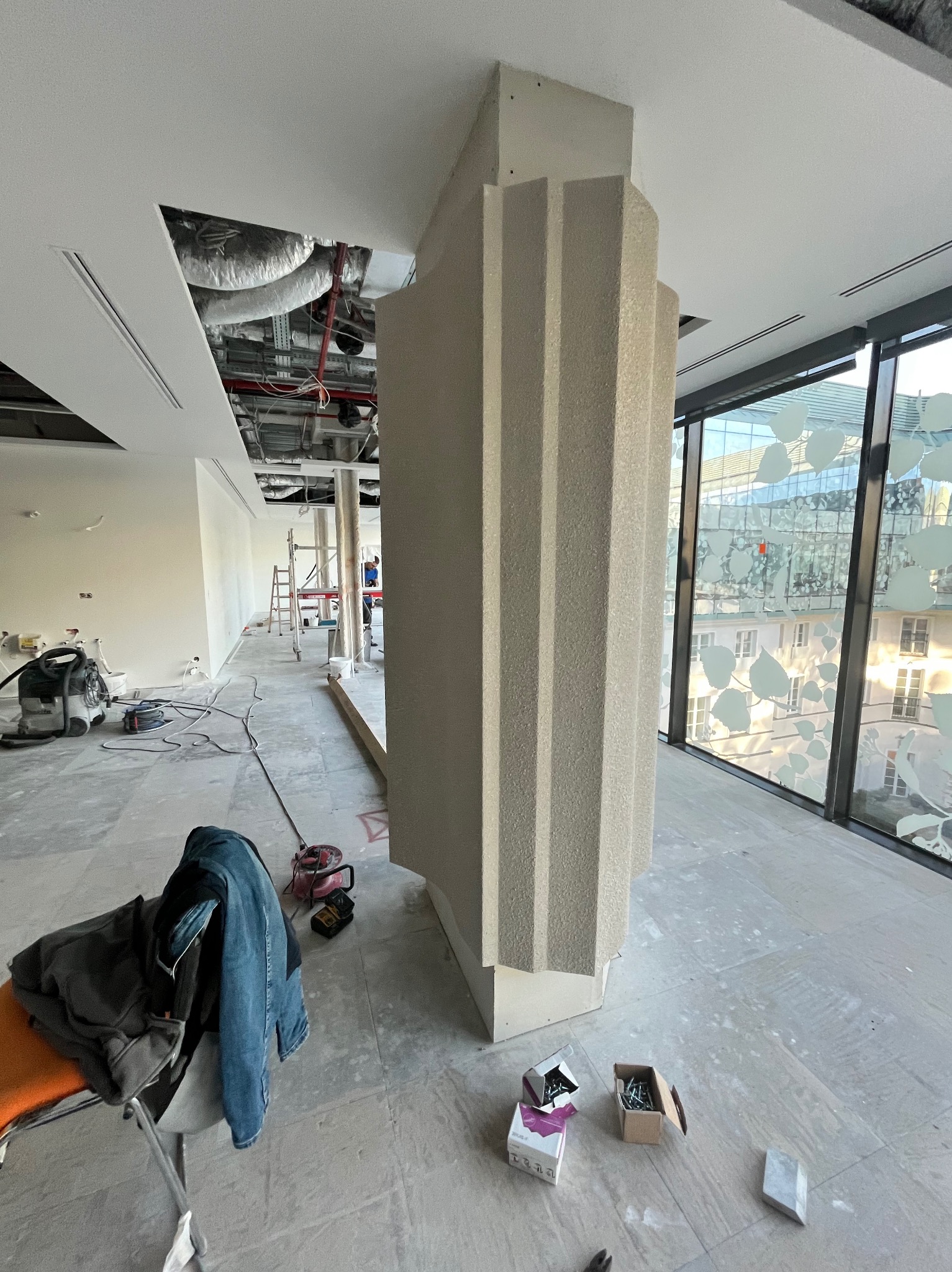 Wnętrze biura w trakcie prac wykończeniowych; kolumna z nałożoną strukturą dekoracyjną, widoczne narzędzia, drabina, instalacje pod sufitem i okna z dekoracyjną folią.