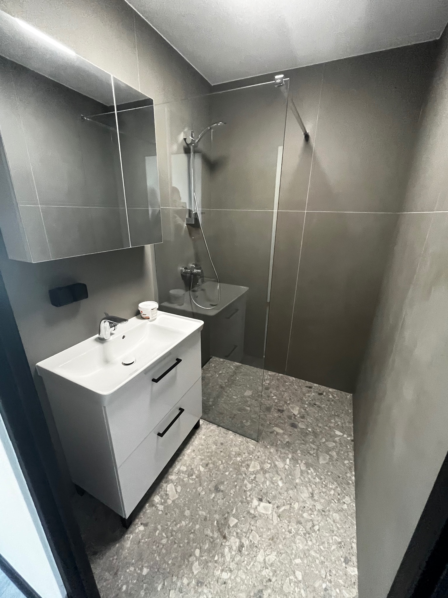 Nowoczesna łazienka z szarymi płytkami, białą szafką pod umywalką z czarnymi uchwytami, szklaną kabiną prysznicową i podłogą z terazzo. Na umywalce stoi biały pojemnik.