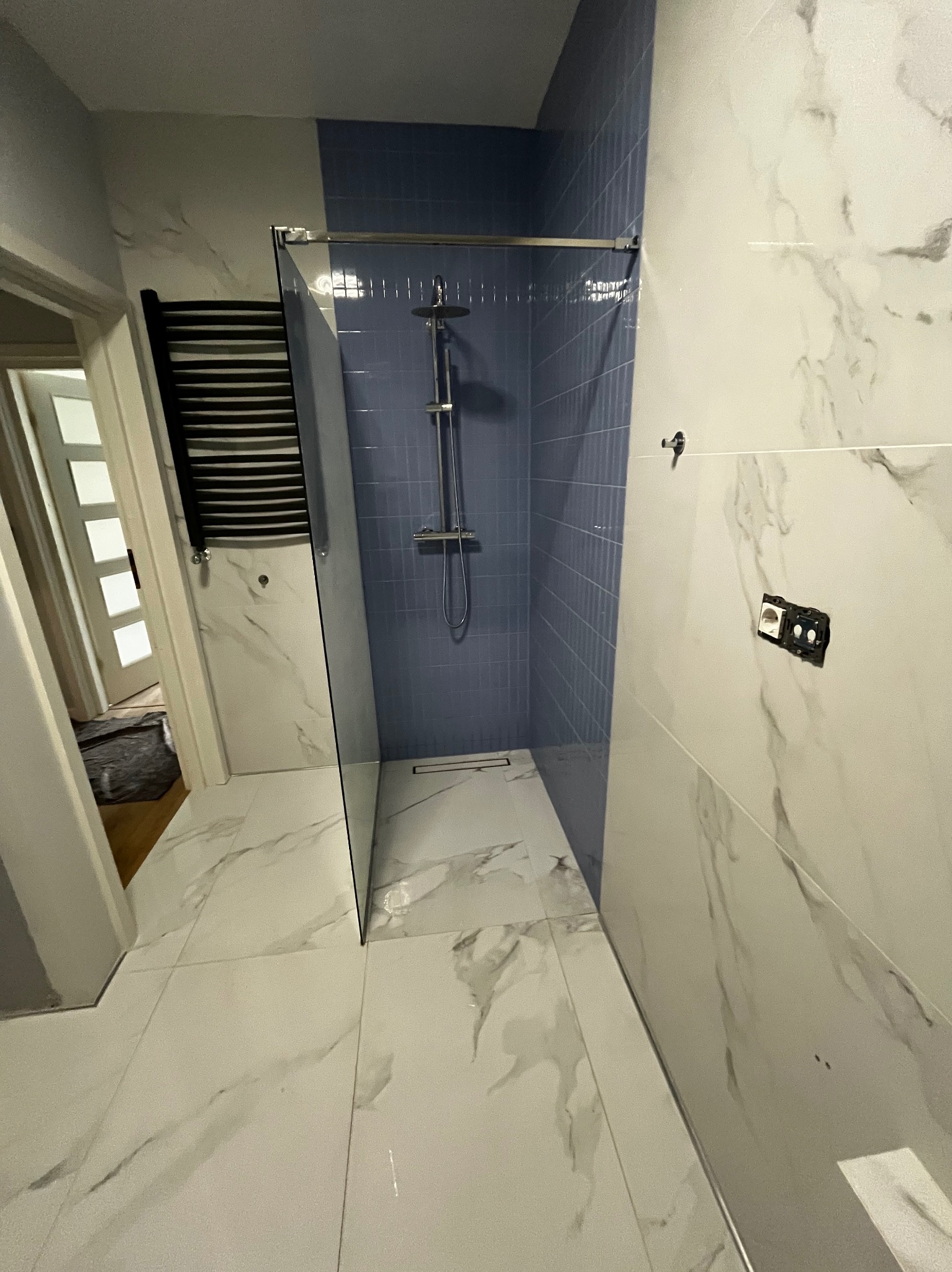 Wykończona łazienka z kabiną prysznicową typu walk-in z niebieskimi płytkami, białe płytki imitujące marmur na ścianach i podłodze, widoczne niedokończone gniazdko elektryczne i grzejnik drabinkowy.