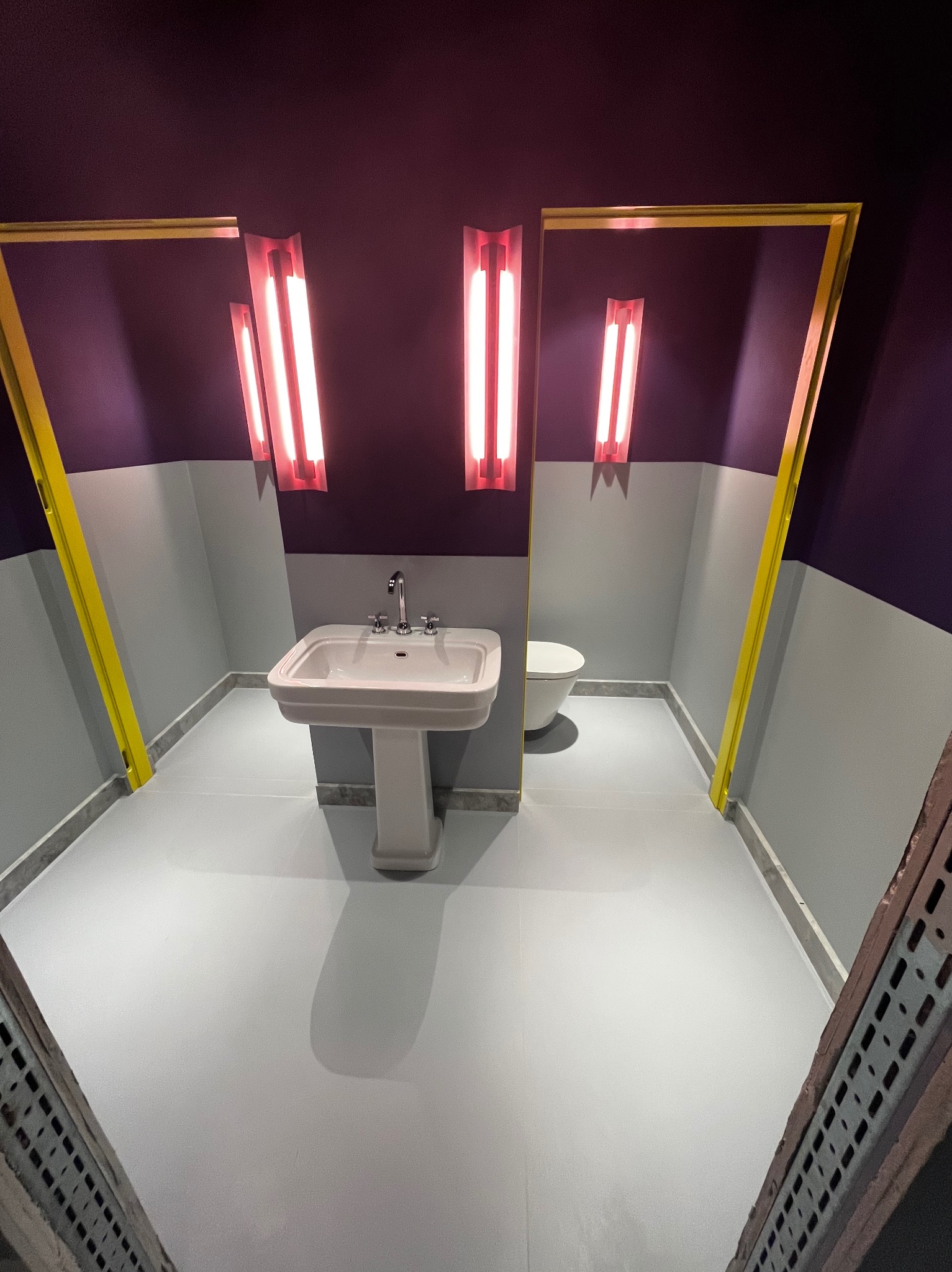 Nowoczesna łazienka w odcieniach szarości i fioletu z geometrycznymi akcentami: umywalka na postumencie, toaleta w tle, podświetlane lustra w złotych ramach i nietypowe oświetlenie neonowe.