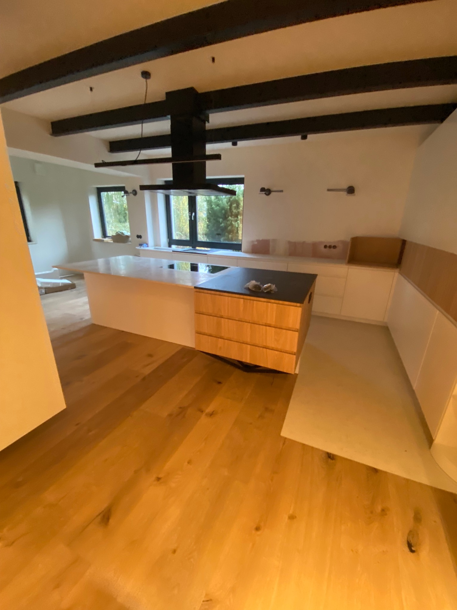 Wnętrze w trakcie wykańczania: drewniana podłoga, białe ściany, zabudowa kuchenna z drewnianymi frontami i czarnym blatem, widoczne czarne belki stropowe i okna z widokiem na zieleń.