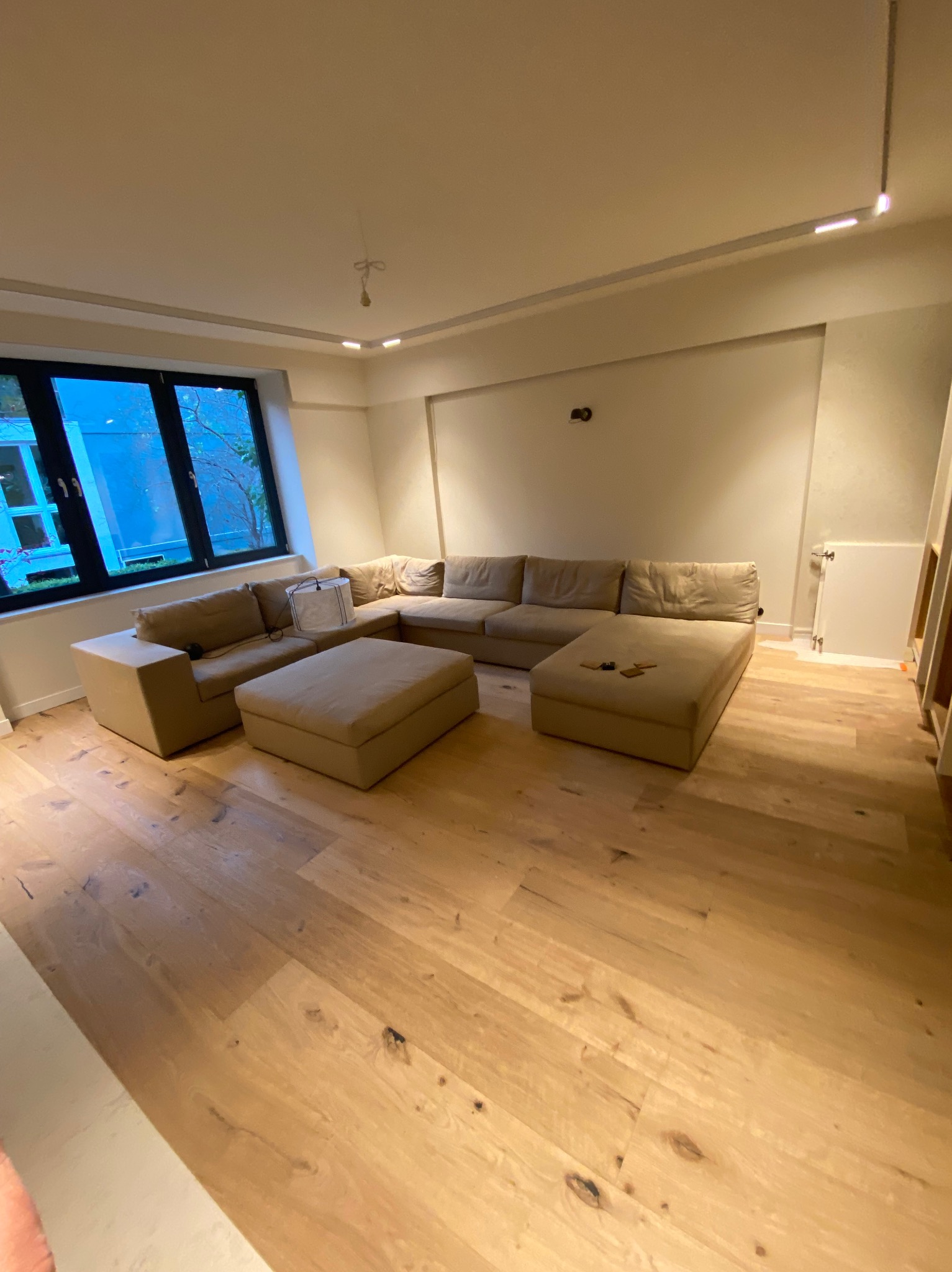 Jasne panele podłogowe z widocznymi słojami, w tle narożna sofa w odcieniach beżu i duże okno z czarną ramą.