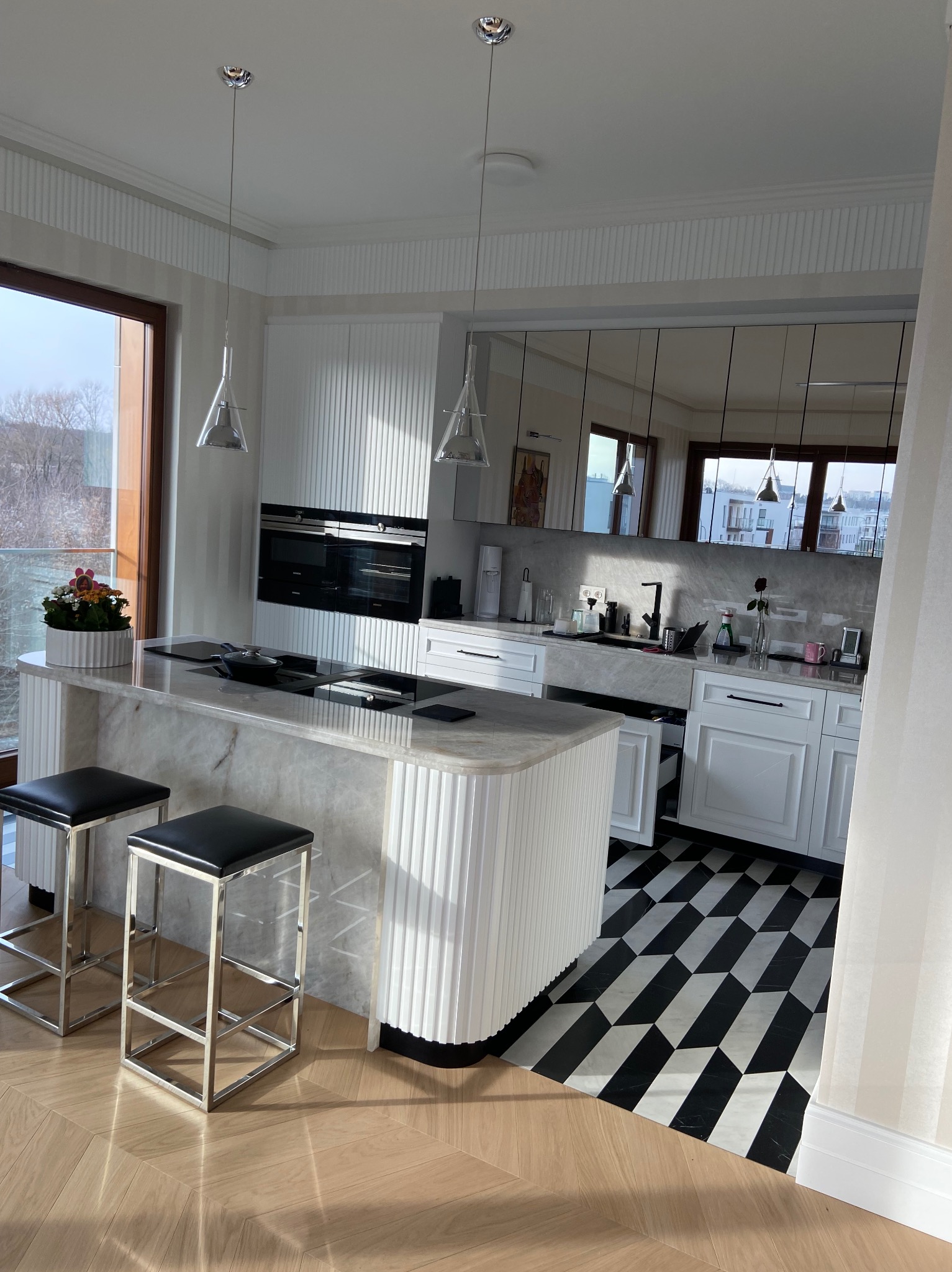 Nowoczesna kuchnia z marmurowym blatem wyspowym, białymi szafkami z pionowymi żłobieniami, czarno-białą podłogą w geometryczny wzór i drewnianą podłogą w jodełkę, oświetlona wiszącymi lampami...