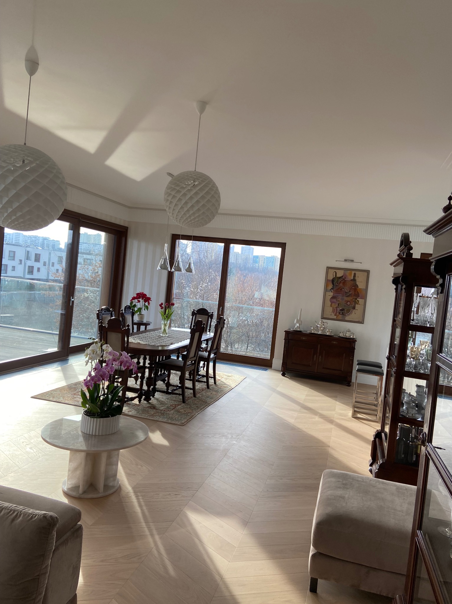 Jasny salon z parkietem w jodełkę, dużymi oknami balkonowymi, okrągłym stołem z krzesłami, kwiatami w wazonach i dekoracyjnymi lampami wiszącymi.