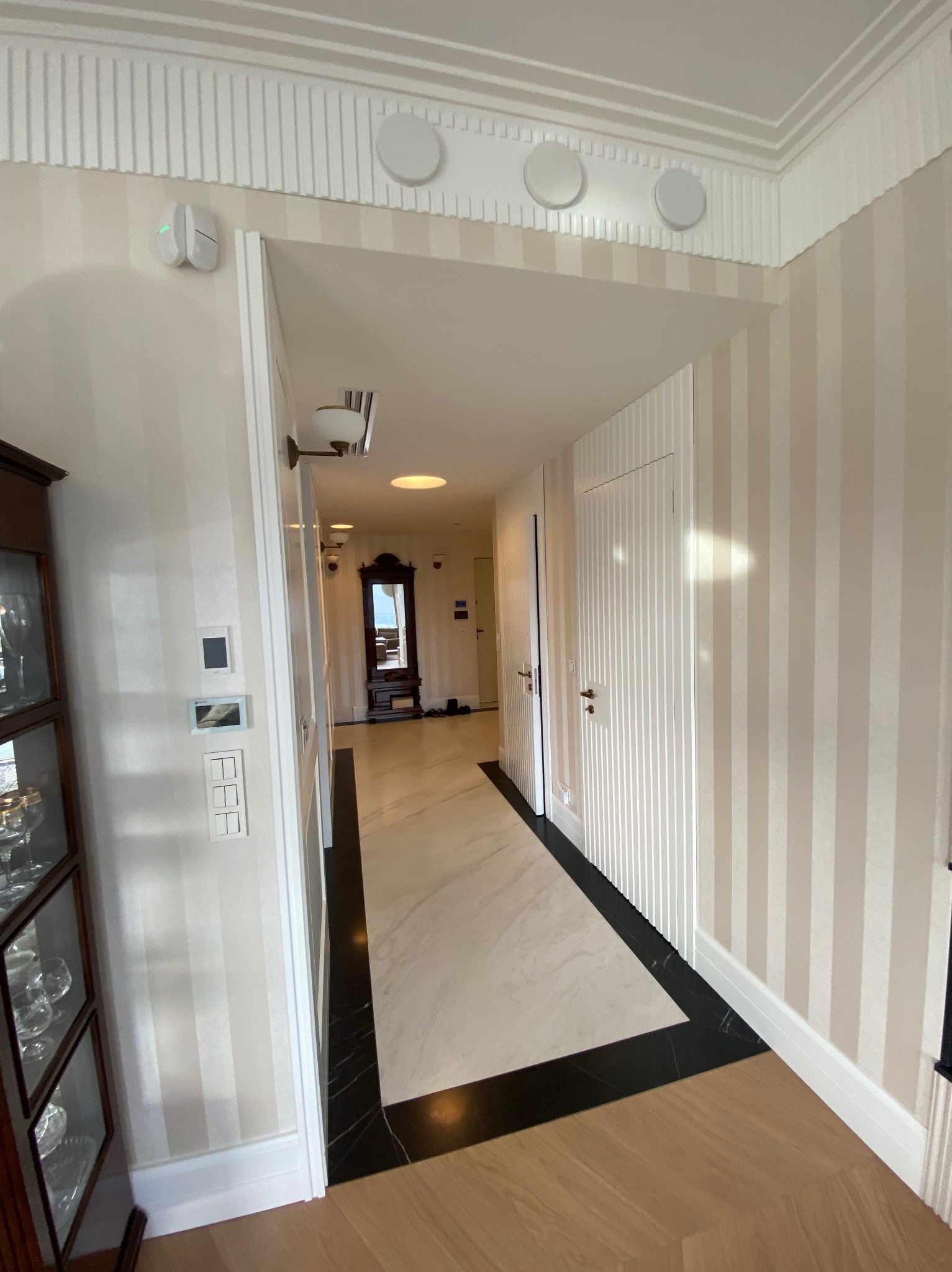Wnętrze korytarza z kremowo-białymi tapetami w pionowe pasy, marmurową posadzką z czarnymi akcentami i drewnianą podłogą, widziane z perspektywy wejścia.