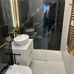 RH-BUD - Elegancka łazienka z czarnymi marmurowymi ścianami, białą umywalką nablatową z marmurowym wzorem, złotymi akcentami w oświetleniu i grzejniku, oraz toaletą wiszącą.