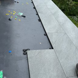 RH-BUD - Układanie szarych płytek na tarasie, widoczne elementy dystansowe i podkładki. Częściowo ułożona podłoga, w tle zieleń drzew. Prace glazurnicze na zewnątrz.