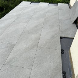 RH-BUD - Montaż szarych, prostokątnych płyt tarasowych na regulowanych wspornikach. Widoczne elementy hydroizolacji. Tło: zieleń drzew i krzewów.