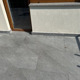 RH-BUD - Wykończony balkon z szarymi płytkami, widoczne detale łączeń. Ściana o jasnej, chropowatej fakturze. Brązowa rama okienna i cień rzucany przez balustradę.