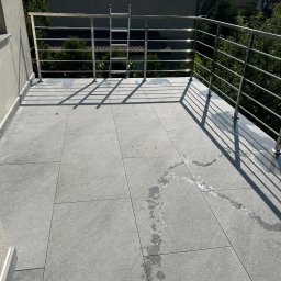 RH-BUD - Wykończony balkon z szarą, dużą płytką i metalową balustradą. Widoczne ślady po myciu płytek. W tle zieleń drzew i zabudowa miejska.