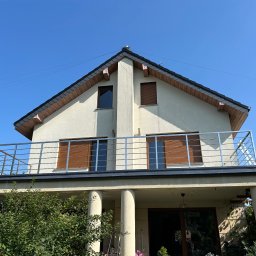 RH-BUD - Dwupiętrowy dom z balkonem i brązowymi okiennicami, widziany z dołu na tle błękitnego nieba. Stalowa balustrada balkonu i kolumny podtrzymujące balkon.