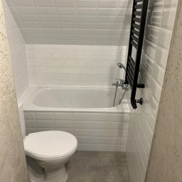 RH-BUD - Biała, wąska łazienka z toaletą, wanną, grzejnikiem drabinkowym i płytkami typu cegiełka. Ujęcie z poziomu podłogi, widoczne detale wykończenia.