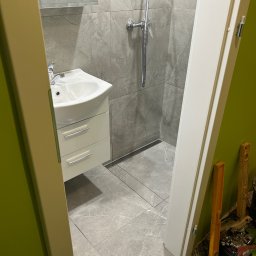 RH-BUD - Wykończona łazienka z szarymi płytkami, kabiną prysznicową i białą szafką z umywalką, widok zza futryny drzwi. Minimalistyczny design, nowoczesne wykończenie.