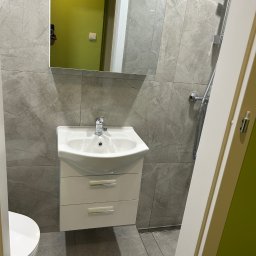 RH-BUD - Wykończona łazienka w szarościach: umywalka z szafką, lustro odbijające zieloną ścianę, szare płytki imitujące marmur, chromowana armatura i kabina prysznicowa.