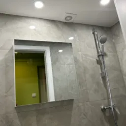 Wykończona łazienka z szarymi płytkami imitującymi marmur, lustrem wbudowanym w ścianę i chromowaną baterią prysznicową. Widoczny fragment wentylacji.