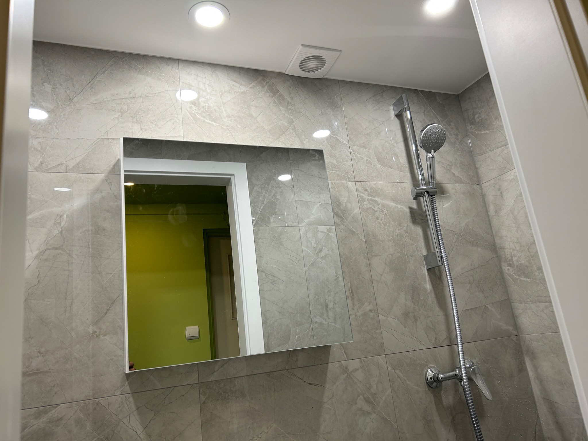 Wykończona łazienka z szarymi płytkami imitującymi marmur, chromowaną armaturą i lustrem, w którym odbija się zielona ściana. Widoczny wentylator sufitowy.