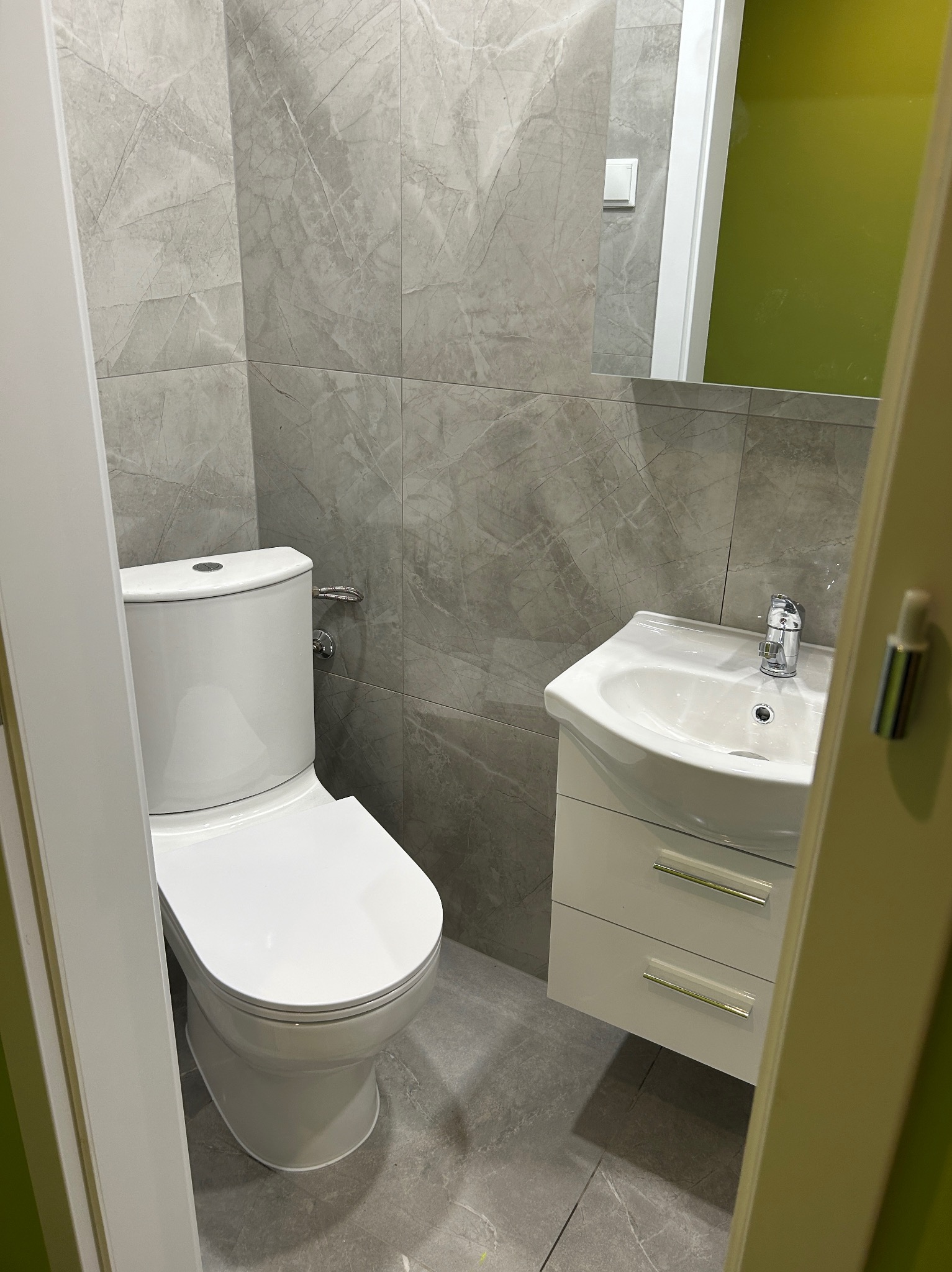 Wykończona łazienka z szarymi płytkami imitującymi marmur, białą toaletą i umywalką z szafką. Widok zza uchylonych drzwi.