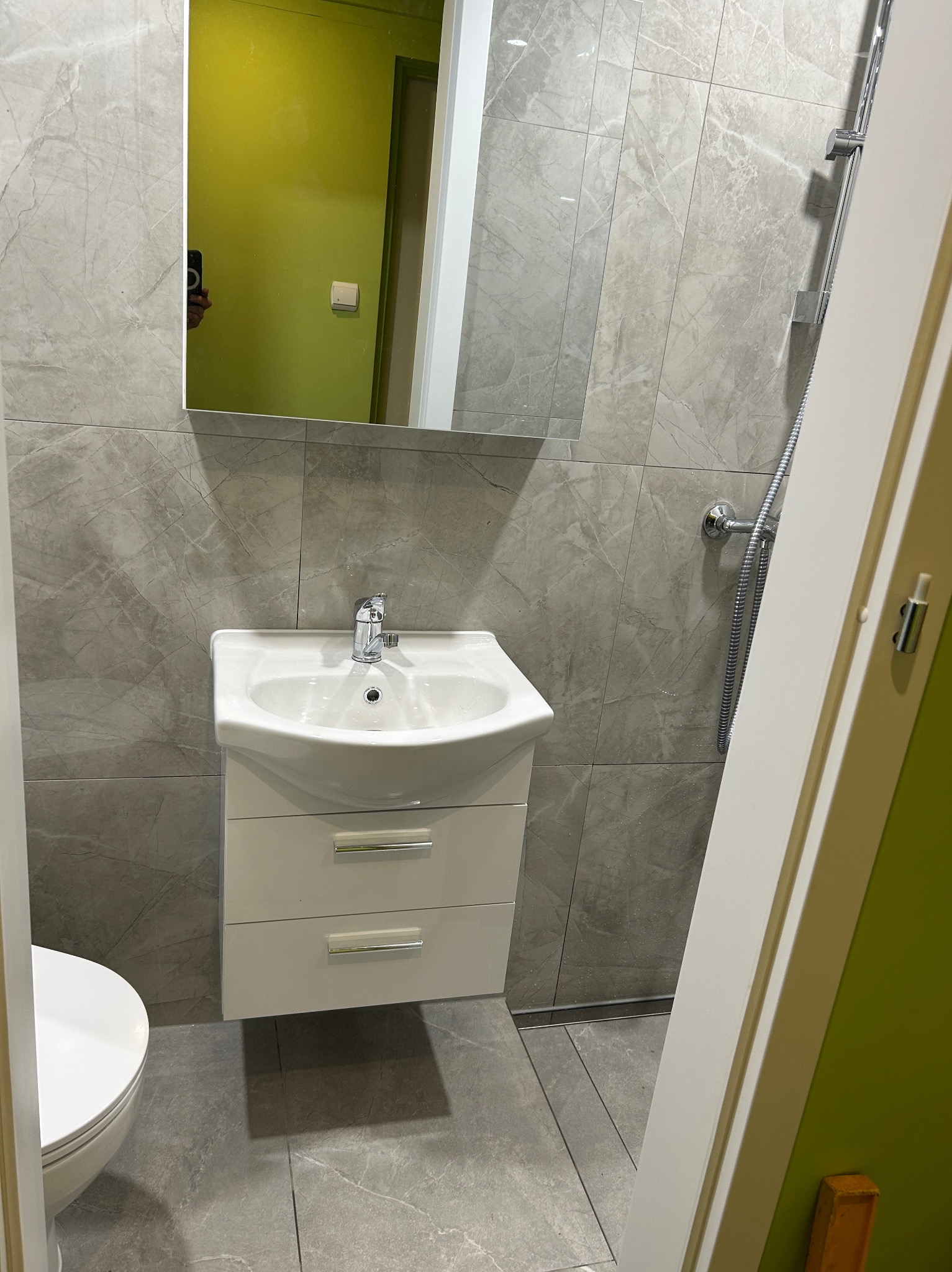 Nowoczesna łazienka z szarymi płytkami imitującymi marmur, białą szafką pod umywalką i chromowaną armaturą. W lustrze odbija się zielona ściana.
