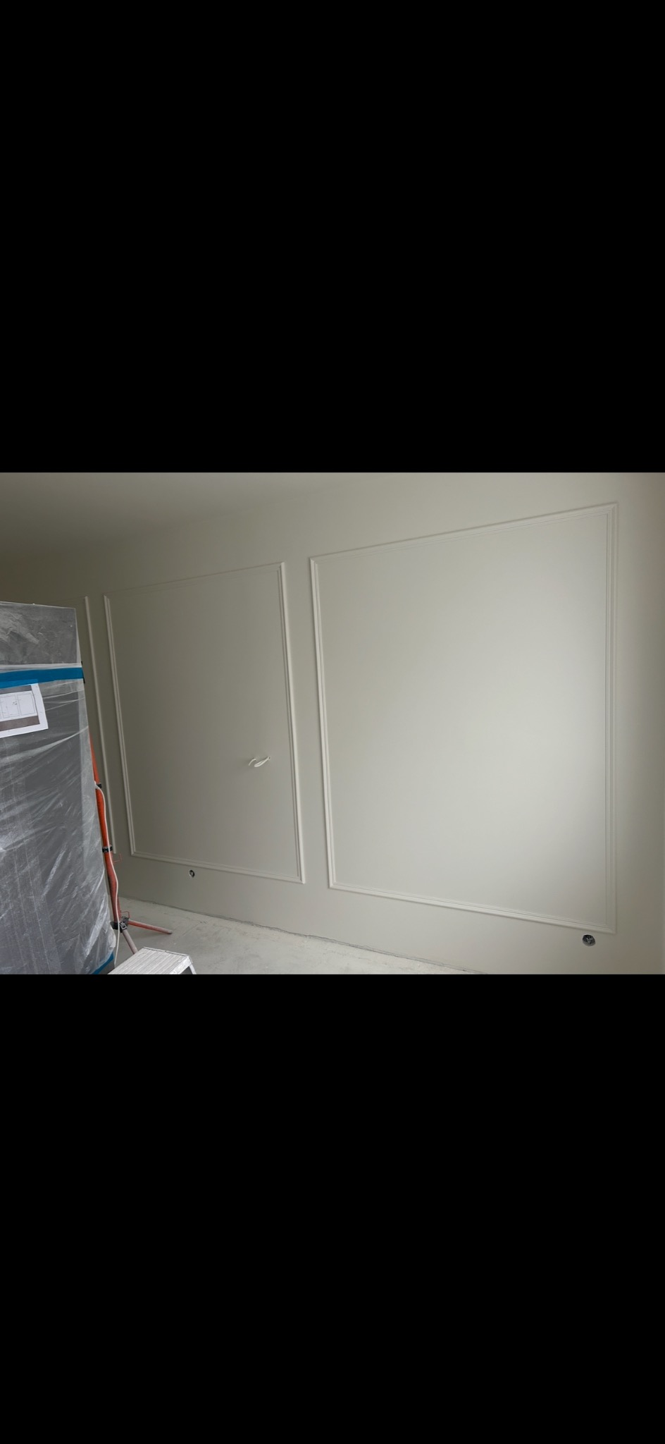 Ściana z dekoracyjnymi panelami w kolorze złamanej bieli. Widoczne delikatne listwy tworzące prostokątne ramy. Częściowo widoczne zabezpieczone folią elementy.