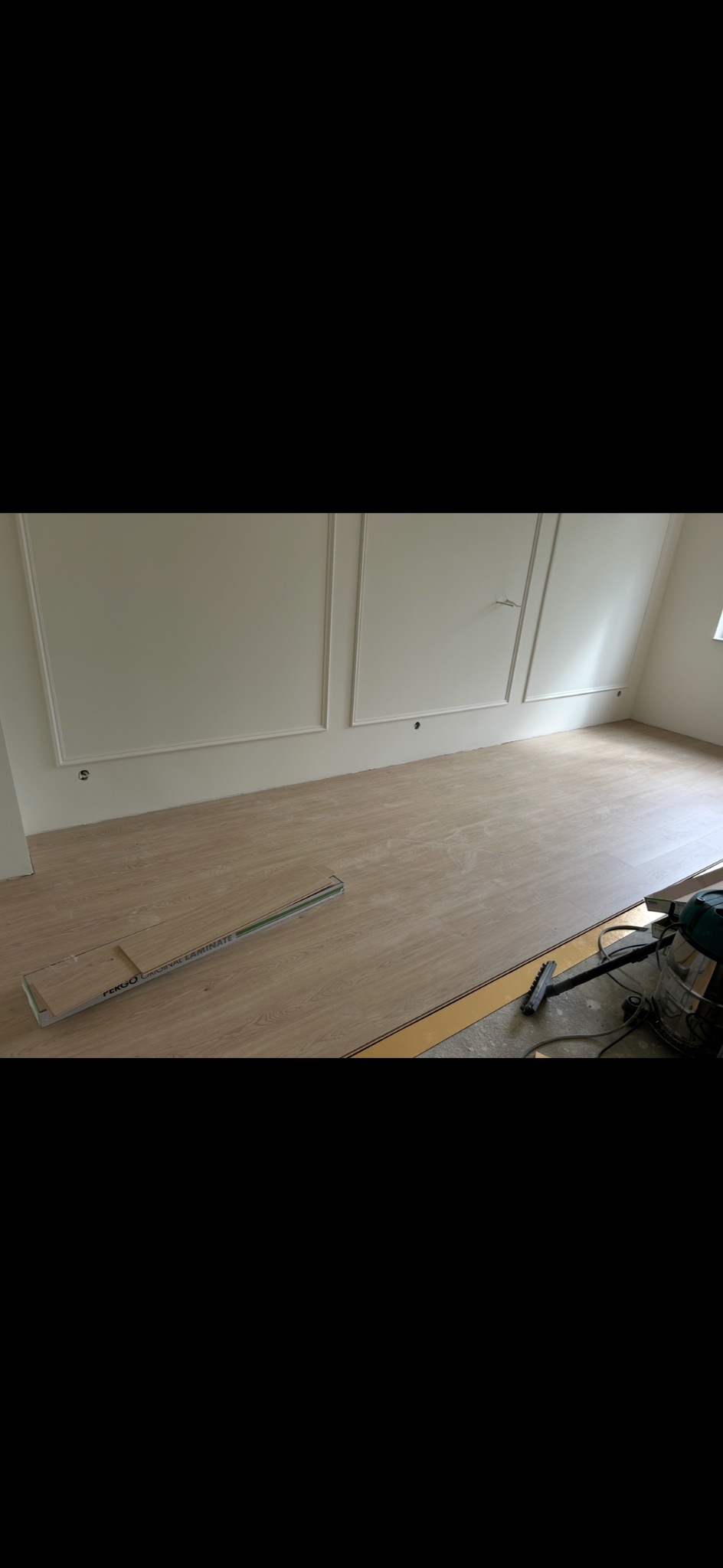 Układanie paneli podłogowych w pokoju z dekoracyjnymi listwami. Widoczna paczka paneli Fergo i odkurzacz. Częściowo ułożona podłoga, jasne panele.