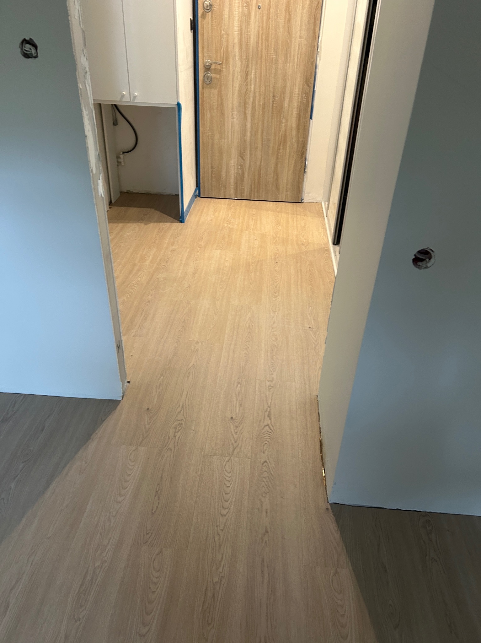 Ukończona podłoga z paneli w jasnym odcieniu drewna, widok z perspektywy wejścia, ściany przygotowane do malowania, fragment szafki z instalacją w tle.