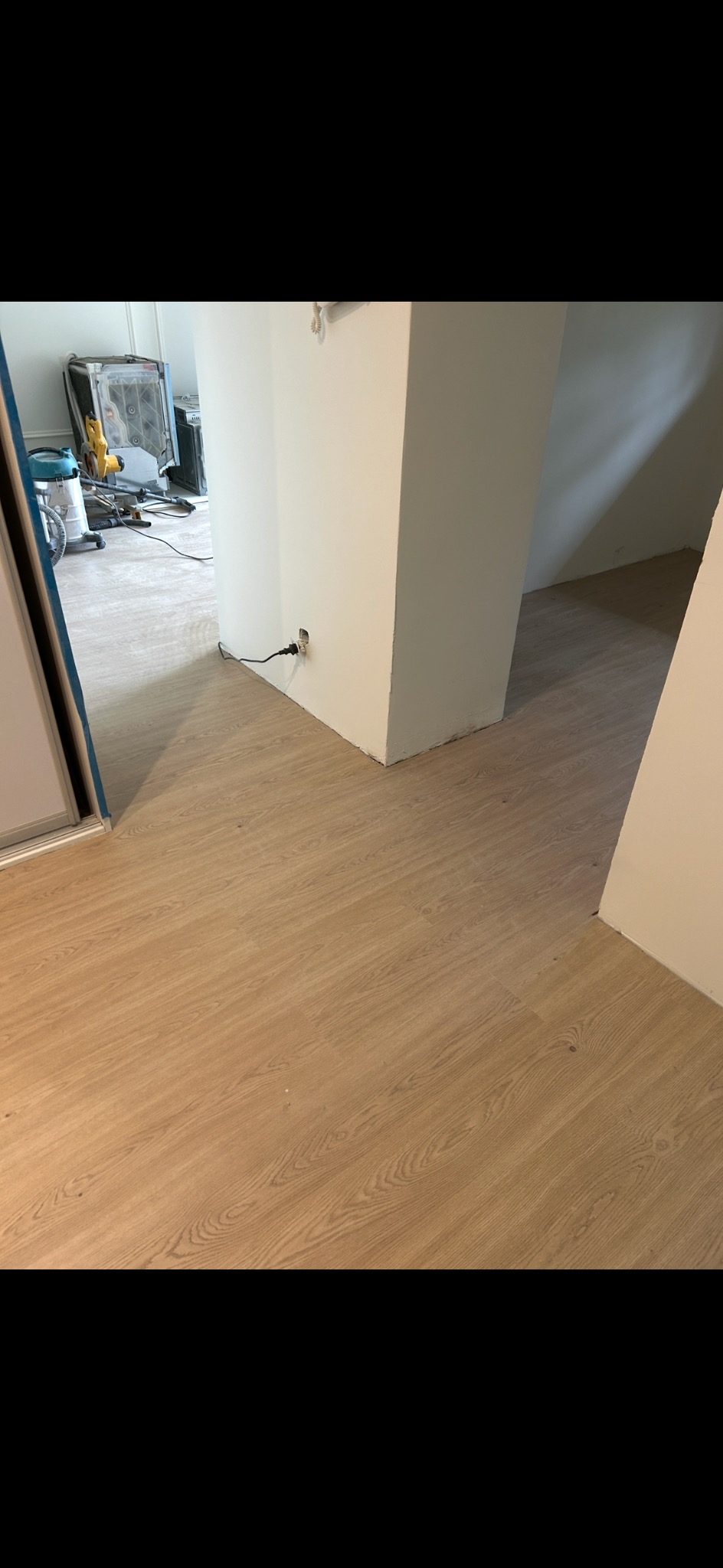 Ułożona podłoga z paneli w jasnym odcieniu drewna, widoczny fragment ściany z gniazdkiem elektrycznym i narzędzia w tle. Układanie paneli podłogowych.