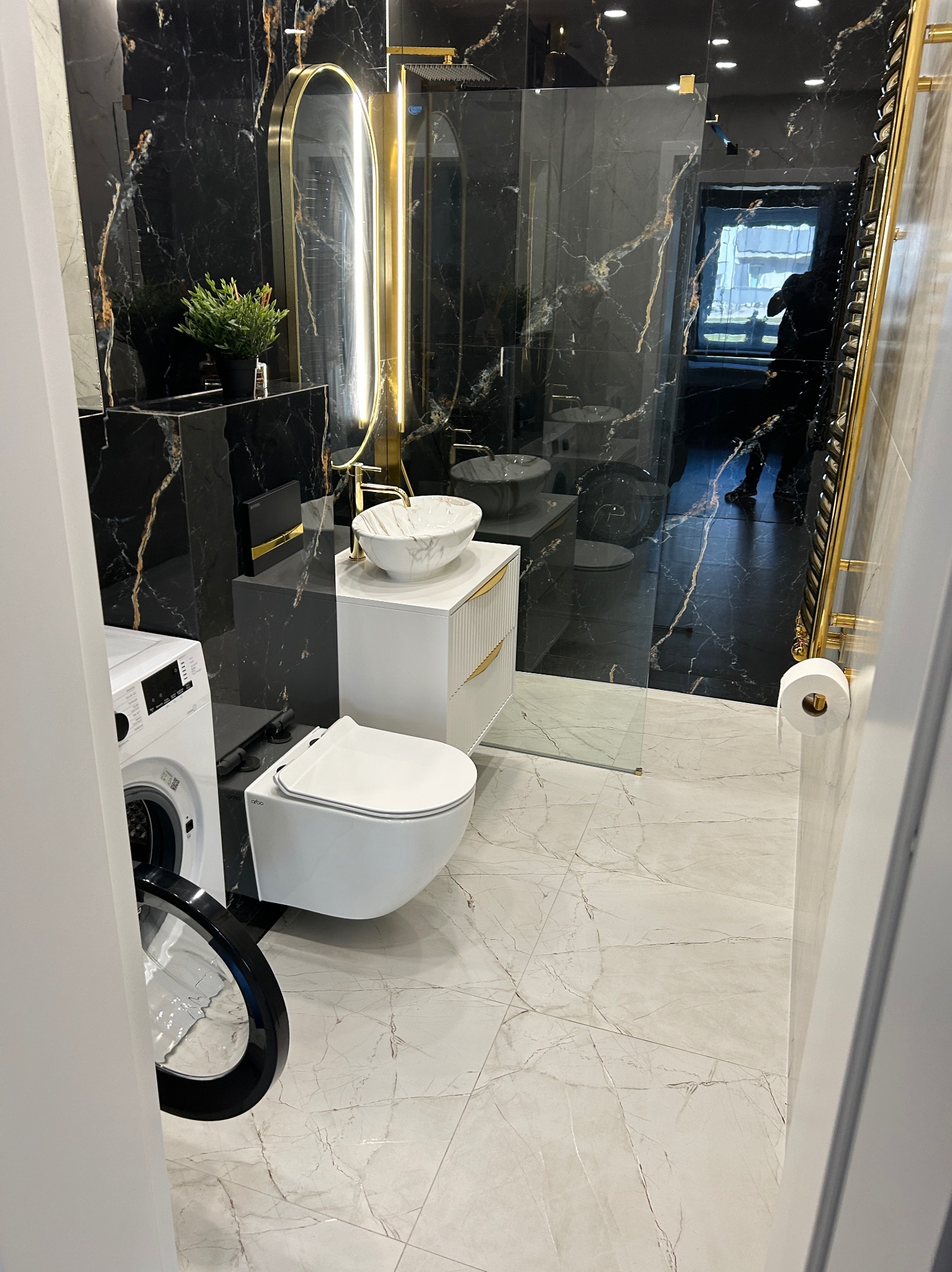Elegancka łazienka z czarnymi marmurowymi ścianami, złotymi akcentami i nowoczesną armaturą. Widoczna pralka z otwartymi drzwiami i marmurowa podłoga.