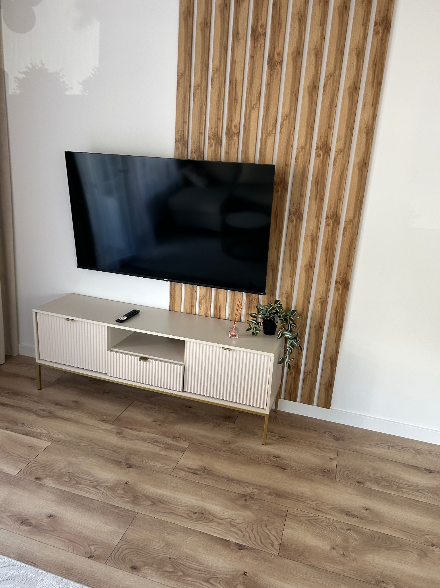 Nowoczesny salon z telewizorem na ścianie z dekoracyjnymi panelami drewnianymi i beżową szafką RTV na złotych nóżkach, uzupełniony o drewnianą podłogę.