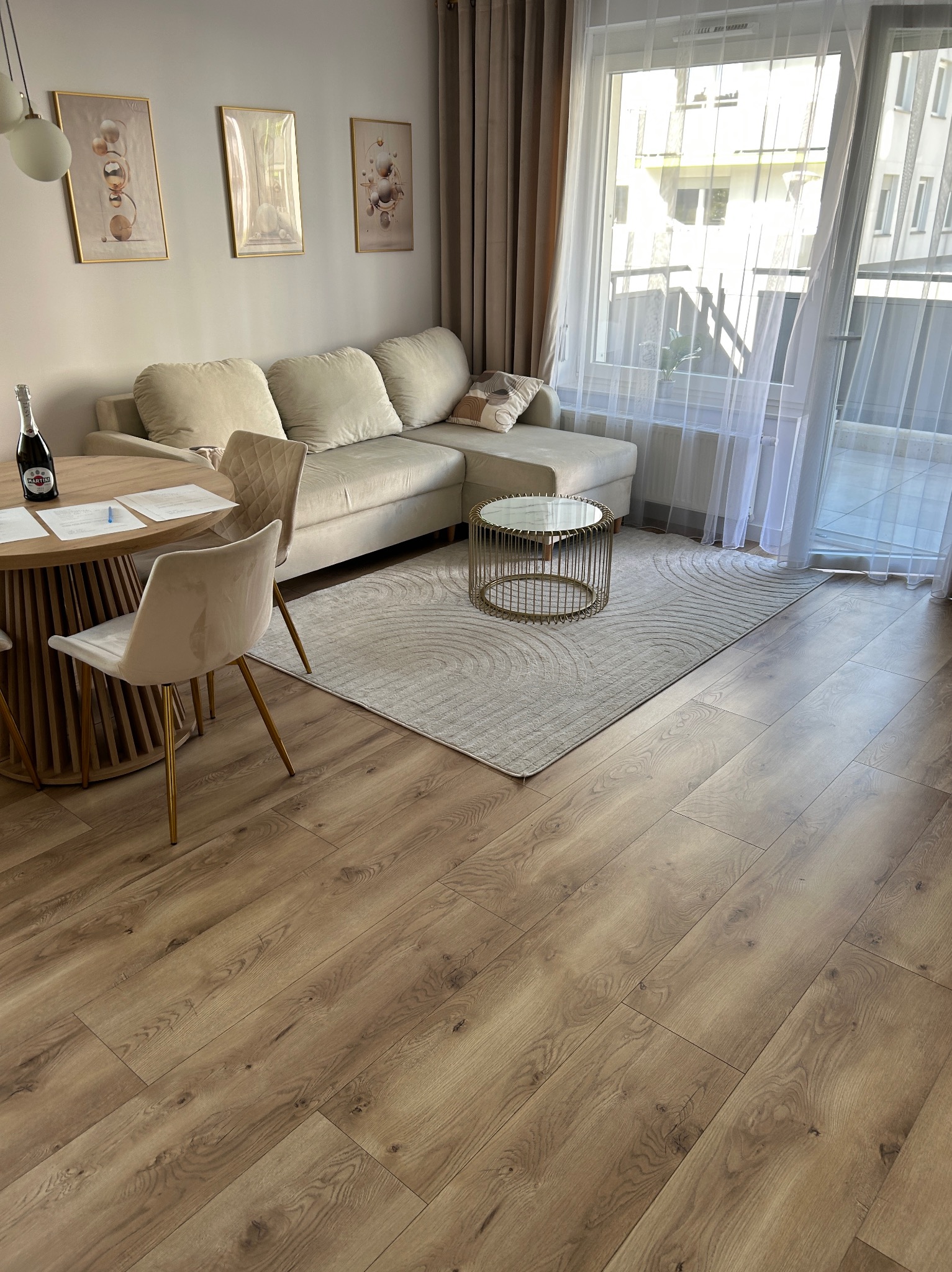 Jasne wnętrze salonu z drewnianą podłogą, beżową sofą, okrągłym stolikiem i dywanem w geometryczne wzory. Na ścianie wiszą trzy obrazy w złotych ramach.