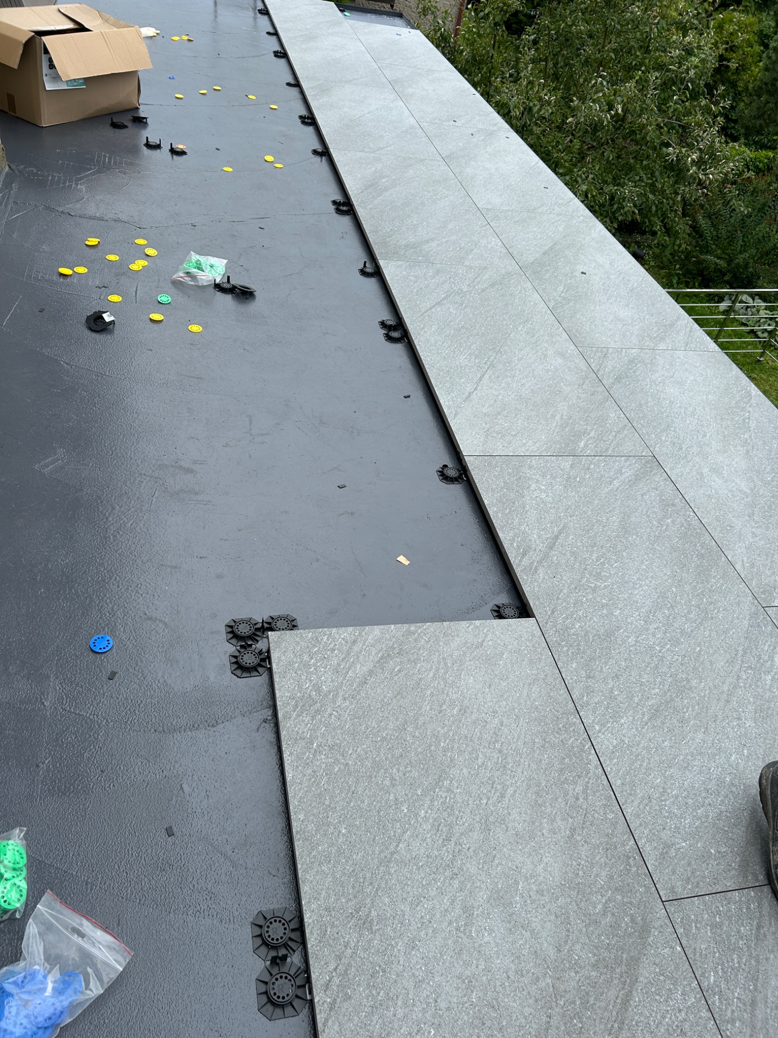 Układanie szarych płytek na tarasie, widoczne elementy dystansowe i podkładki. Częściowo ułożona podłoga, w tle zieleń drzew. Prace glazurnicze na zewnątrz.