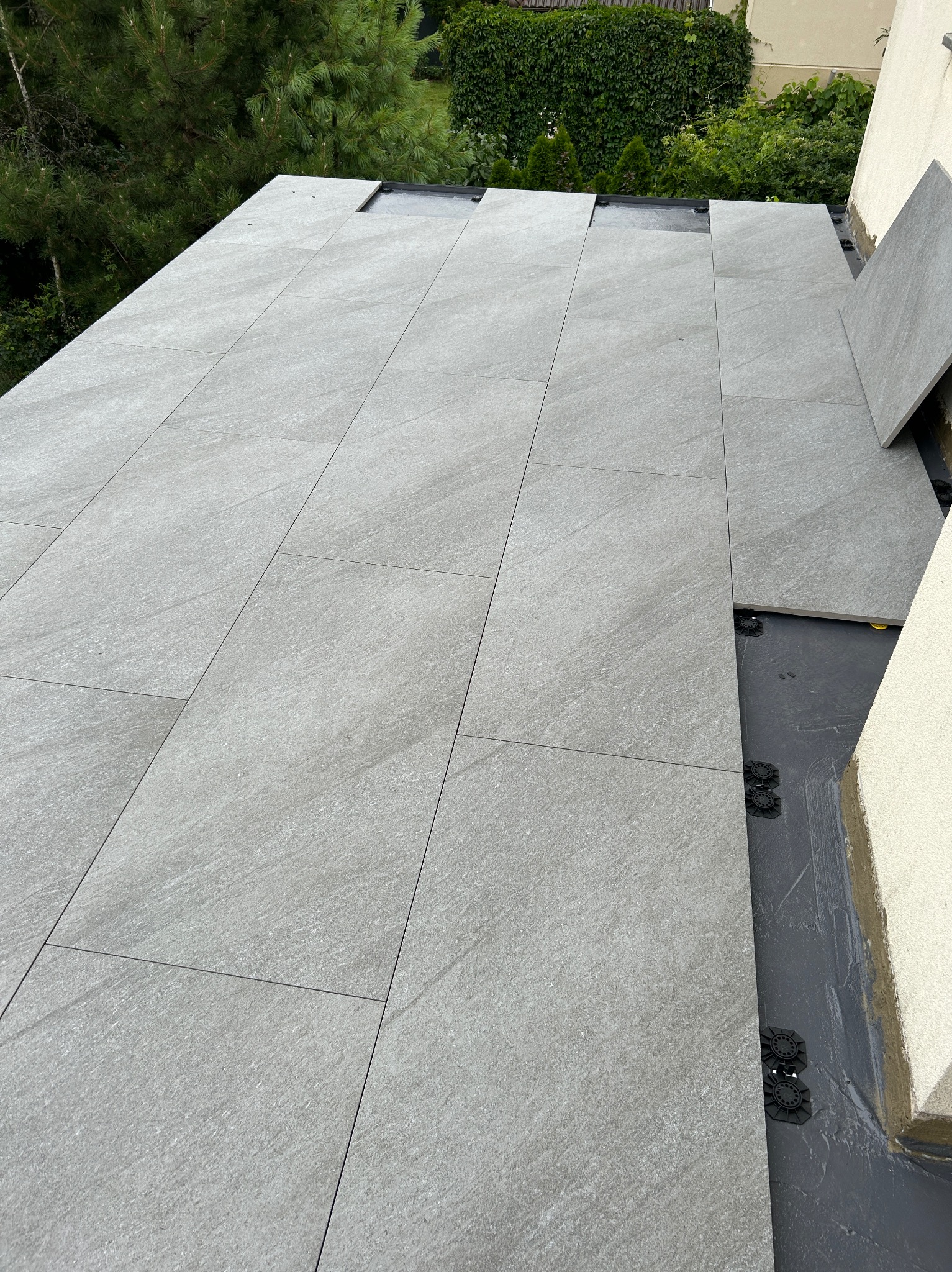 Montaż szarych, prostokątnych płyt tarasowych na regulowanych wspornikach. Widoczne elementy hydroizolacji. Tło: zieleń drzew i krzewów.