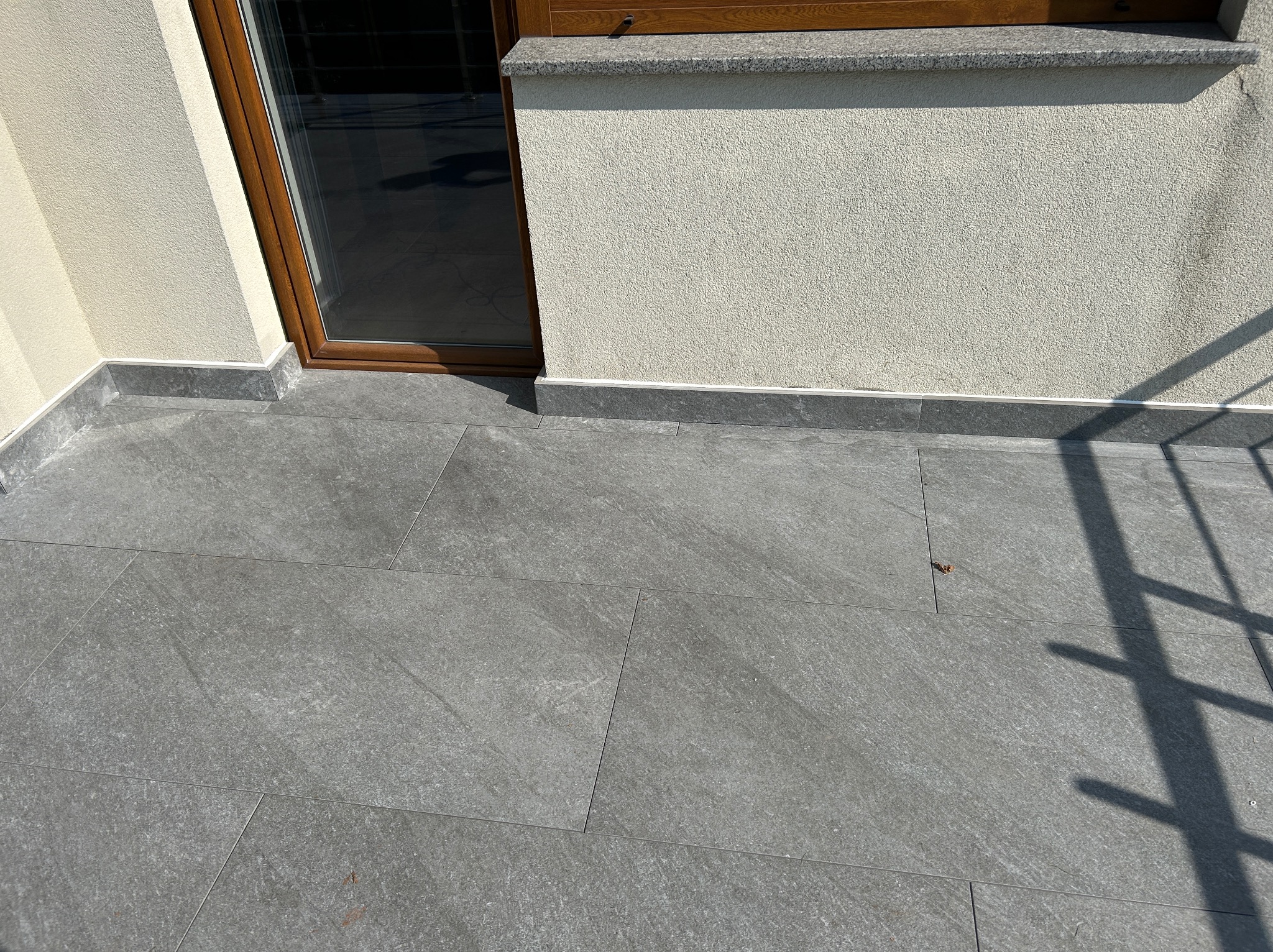 Wykończony balkon z szarymi płytkami, widoczne detale łączeń. Ściana o jasnej, chropowatej fakturze. Brązowa rama okienna i cień rzucany przez balustradę.